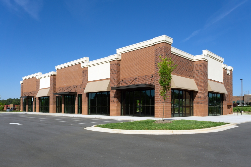 Exterior of a modern commercial strip mall building with a red brick facade, beige awnings, and an empty parking lot in front.