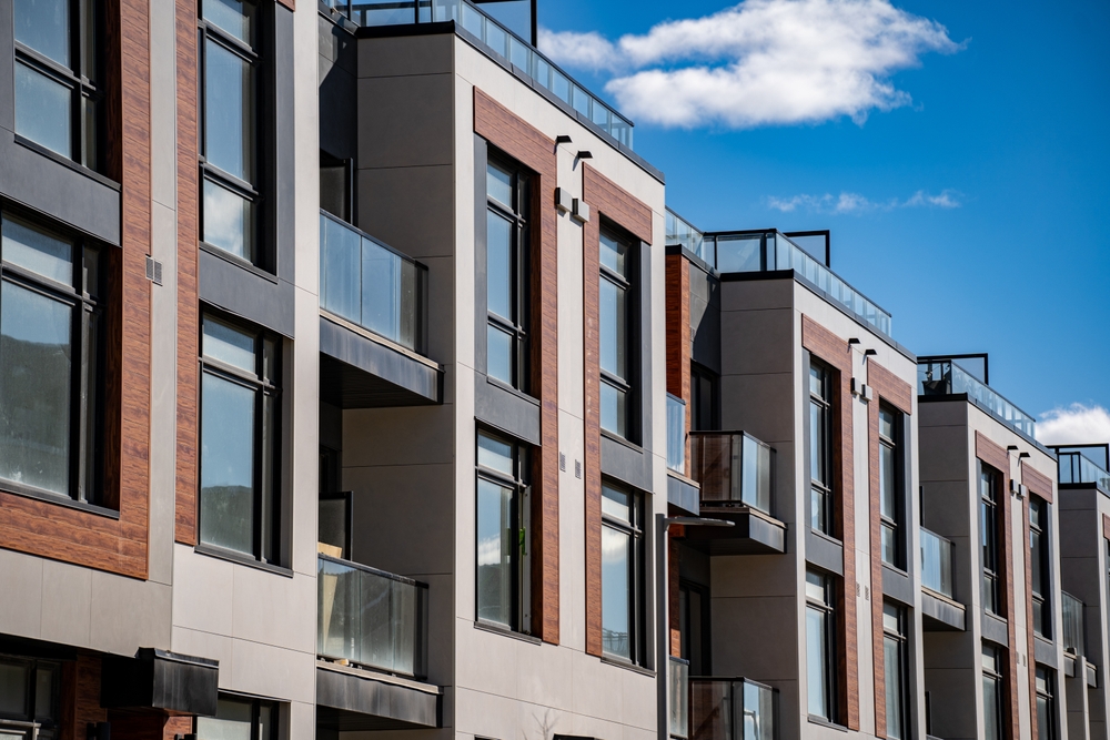 Exterior facade of a modern multi-story apartment complex featuring glass balconies and wood-panel accents against a blue sky.